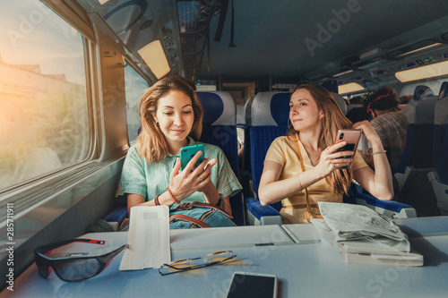 Obraz na płótnie Happy two female friends travelling by long distant high speed train using their smartphones w salonie