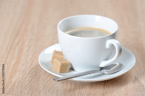 Coffee in a white cup and saucer. On a saucer are two pieces of cane sugar and a teaspoon. Light background. Free space for text.