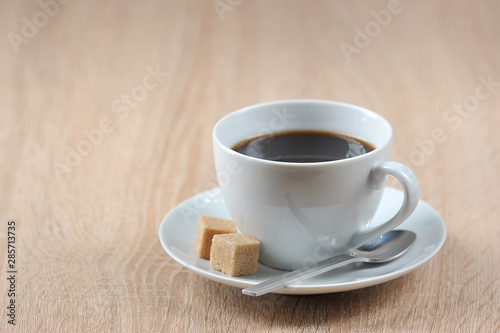 Black coffee in a white cup and saucer. On a saucer are two pieces of cane sugar and a teaspoon. Light background. Free space for text.