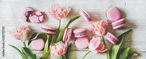 Flat-lay of sweet pink macaron cookies and fresh spring tulip flowers over white wooden background, top view, wide composition. Food texture, background and wallpaper