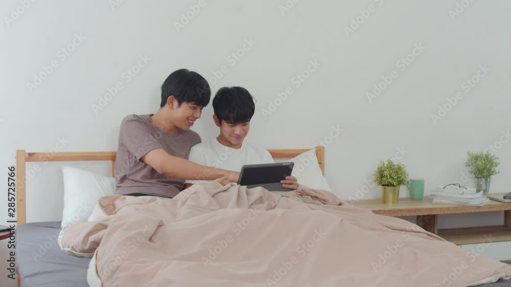 Asian Gay couple using tablet at home. Young Asian LGBTQ men happy relax rest together after wake up, check mail and social media lying on bed in bedroom at home in the morning concept. Asian Gay couple using tablet at home. Young Asian LGBTQ men happy relax rest together after wake up, check mail and social media lying on bed in bedroom at home in the morning concept.