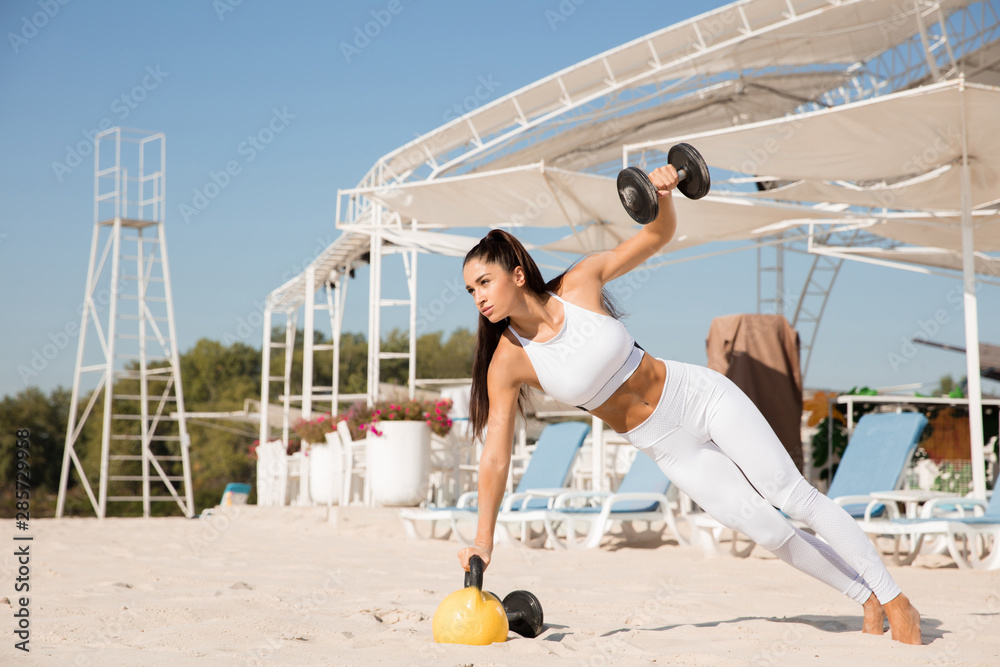 Young healthy woman training upper body with weights at the beach. Single caucasian female model practicing at the river side in sunny day. Concept of healthy lifestyle, sport, fitness, bodybuilding.