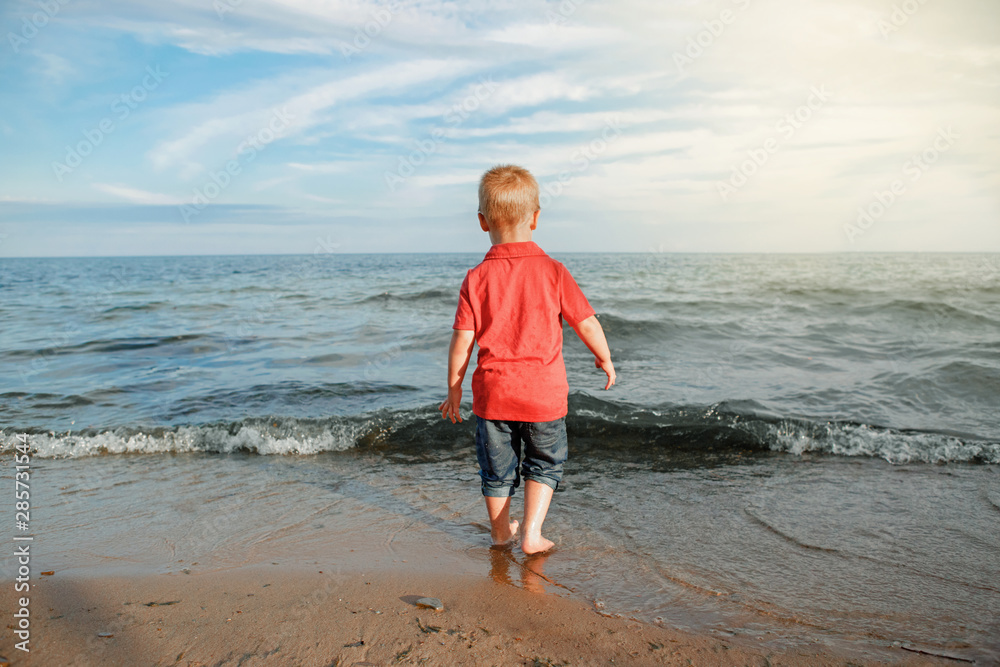 Red-haired toddler child kid in red t-shirt and jeans standing near water on lake sea ocean beach at evening sunset looking far away.  Happy lifestyle childhood concept. View from back.
