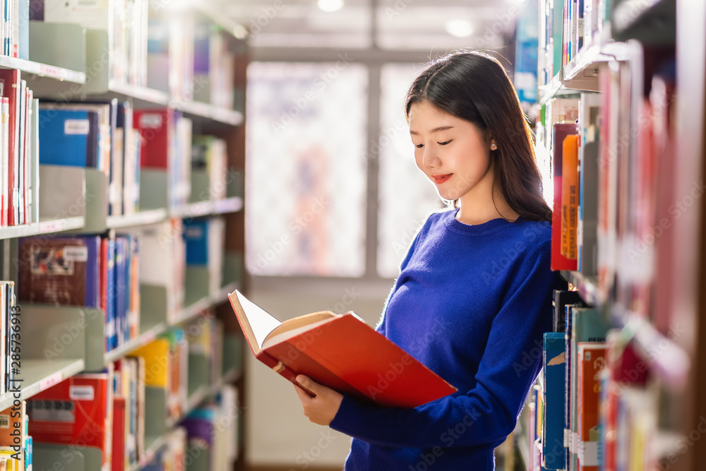 Asian young Student in casual suit standing and reading the book at book shelf in library of university or colleage with various book background, Back to school concept