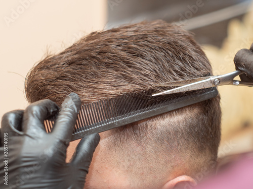 Males hair cutting with comb and scissors, close up view. Hairstylists hands in black rubber gloves cutting hair with comb and scissors. Selective soft focus. Blurred background