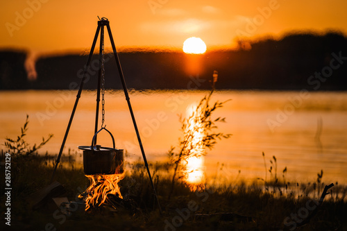 Travel a kettle over a fire burning on the river and sunset background