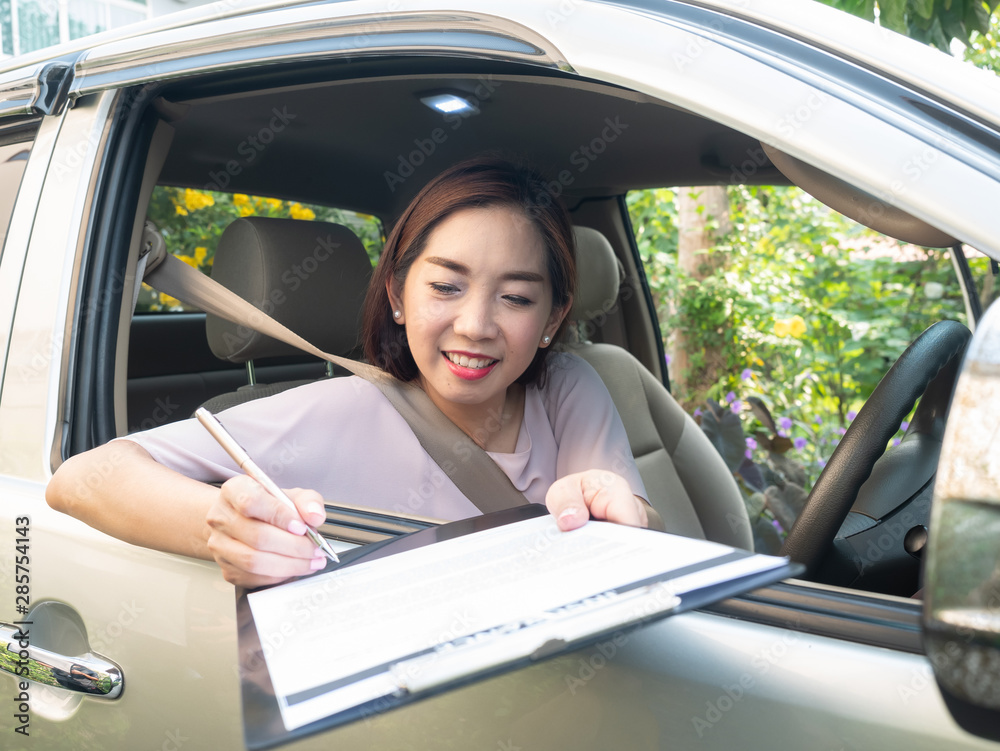 Asian woman inside car reading and signing insurance document.