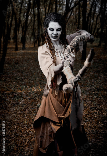 Halloween theme: crazy gloomy voodoo witch killing by stick. Portrait of evil hag in dark forest. Zombie woman (undead)