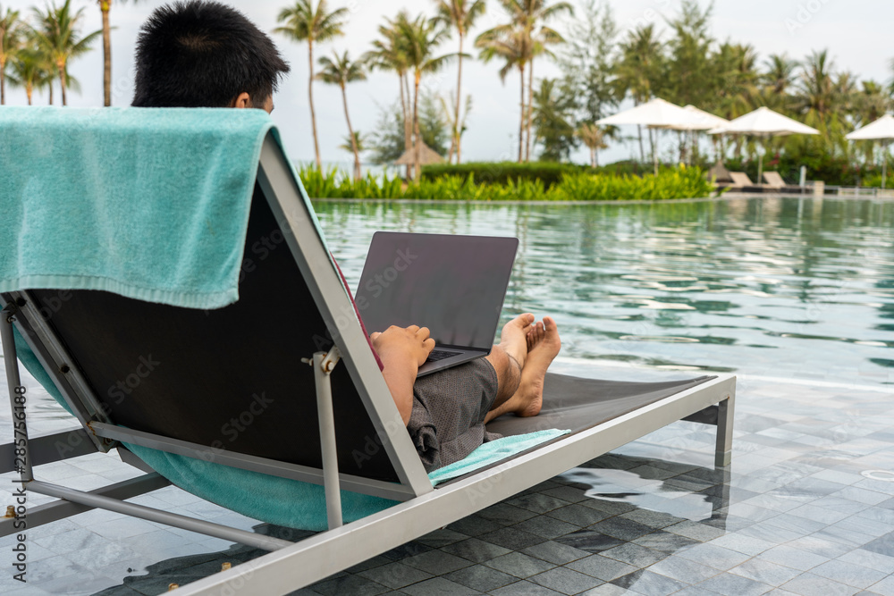Asian man using laptop at swimming pool on summer holiday