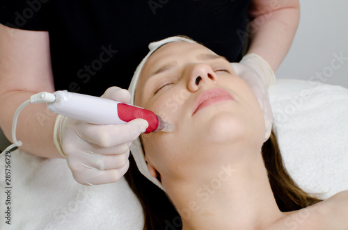 Cosmetologist,beautician in white gloves applying facial dermapen treatment on face of young girl customer in beauty salon.Cosmetology and professional skin care