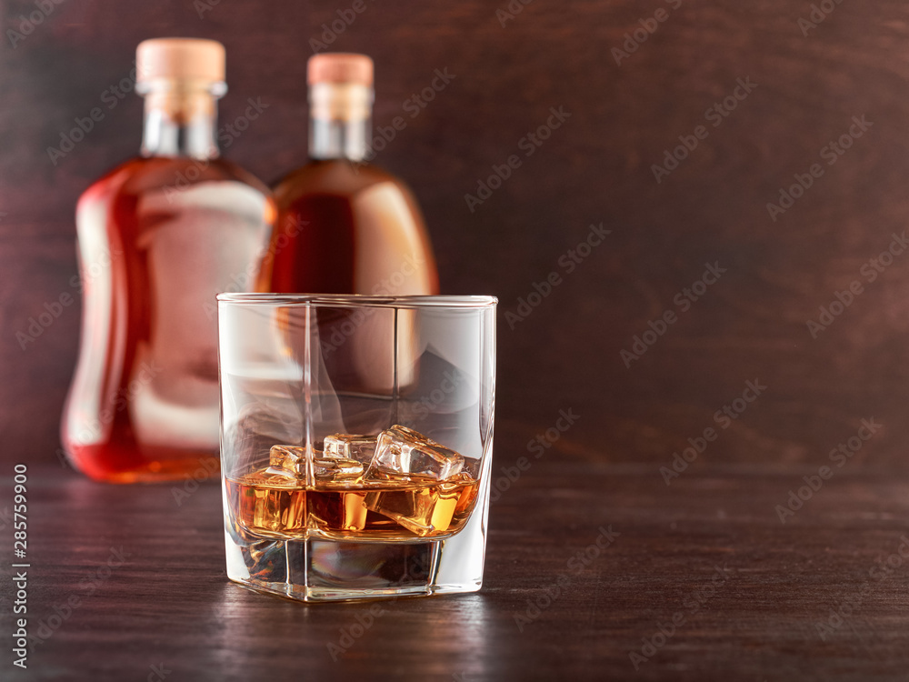 Glass of whiskey with ice on a wooden table, in the background two bottles of whiskey of different shapes Glass of whiskey with ice on a wooden table, in the background two bottles of whiskey of different shapes