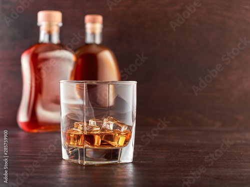 Glass of whiskey with ice on a wooden table, in the background two bottles of whiskey of different shapes