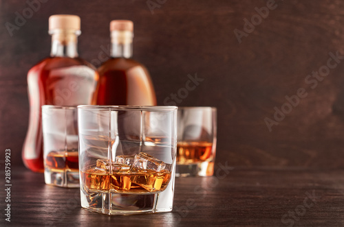 Two glasses of whiskey with ice on a wooden table, in the background two bottles of whiskey of different shapes and a glass of whiskey