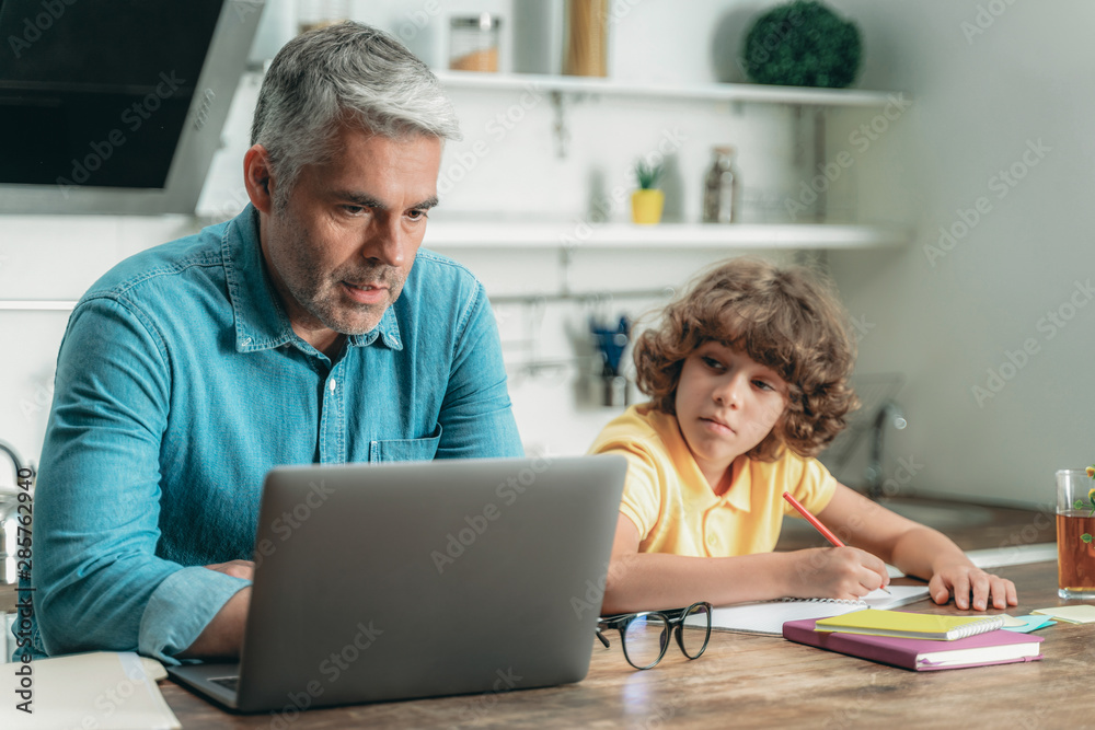 Father using laptop while son making homework Father using laptop while son making homework