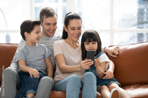 Happy parents with cute kids sit on sofa using phone
