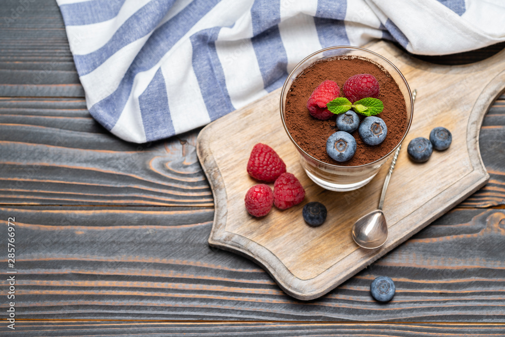 Classic tiramisu dessert portion with blueberries and strawberries in a glass cup on cutting board on wooden background Classic tiramisu dessert portion with blueberries and strawberries in a glass cup on cutting board on wooden background