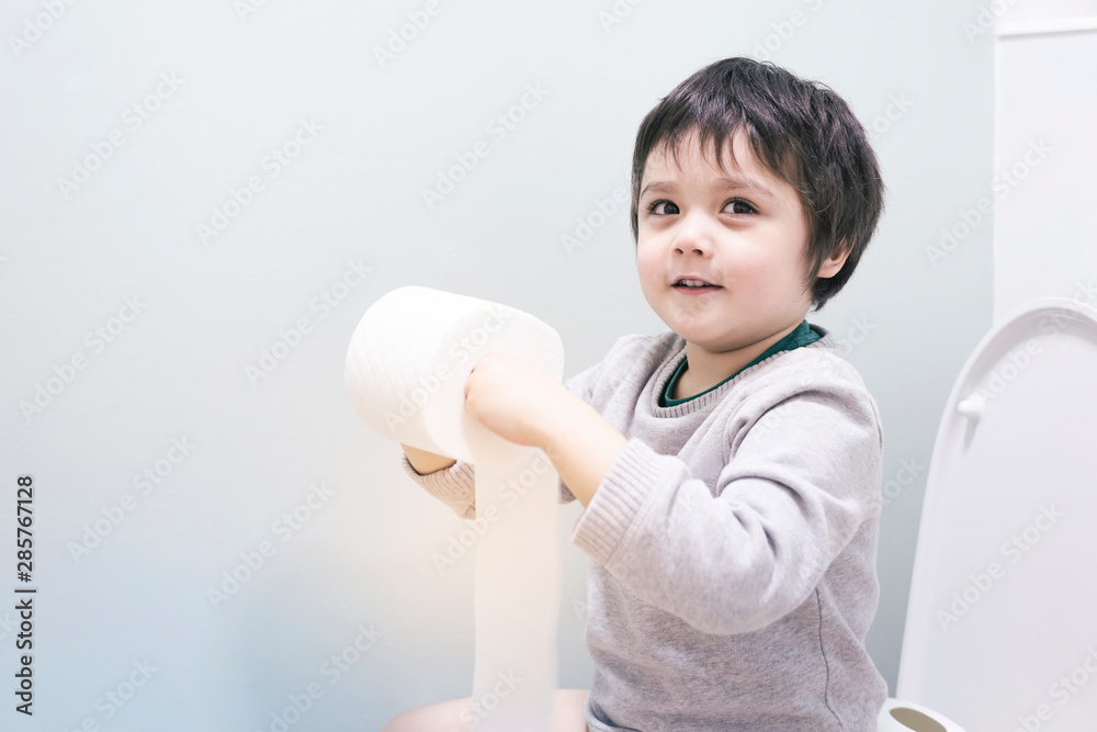Happy boy playing with toilet paper in toilet, Kid sitting with holding a roll of toilet paper and lookign at camera with smiling face, Adorable Child tear toilet roll against light green background. Happy boy playing with toilet paper in toilet, Kid sitting with holding a roll of toilet paper and lookign at camera with smiling face, Adorable Child tear toilet roll against light green background.