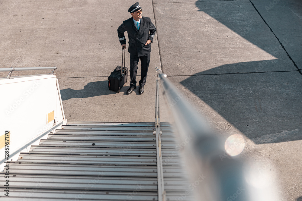 Cheerful adult pilot with suitcase smiling near plane ladder