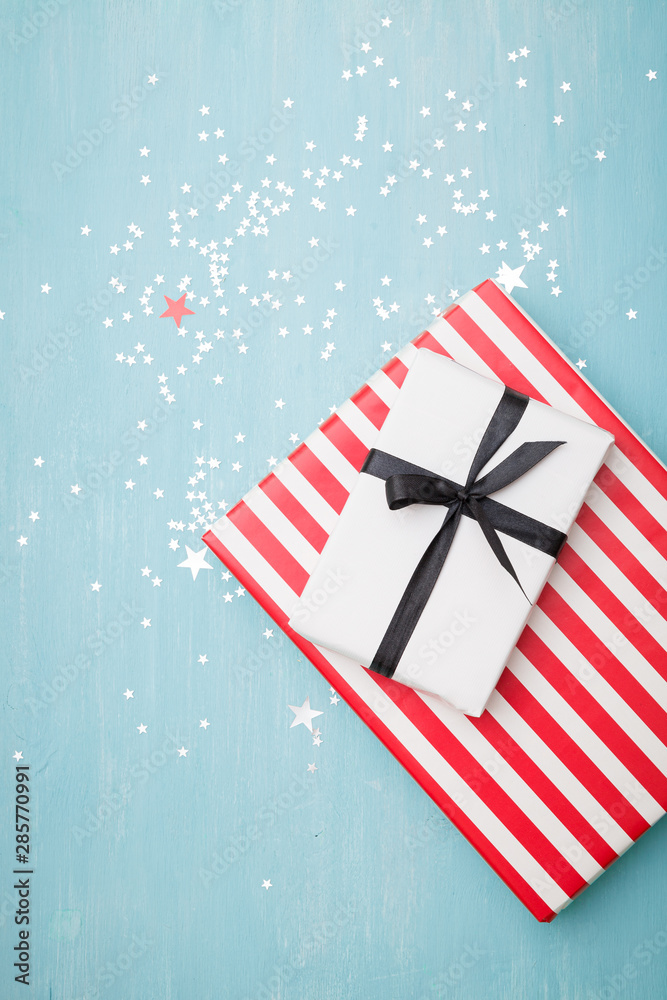 Fototapeta premium Top view of gifts lying on a blue wooden background. Stylish striped gift and a gift wrapped in white paper tied with a black ribbon. Silver stars are scattered on the table like confetti.