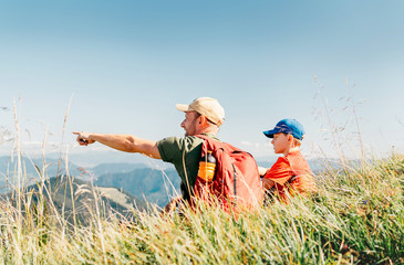 Father showing something interesting to his teenager son sitting on the grass during their mounting hiking walking.