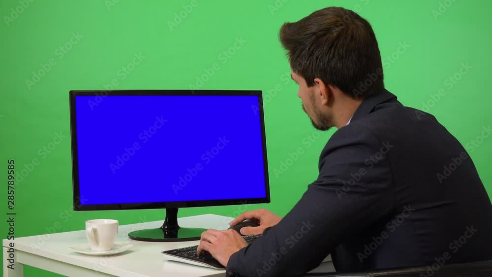 A young businessman sits at a desk and works on a desktop computer with blue screen - green screen studio - he uses mouse and keyboard A young businessman sits at a desk and works on a desktop computer with blue screen - green screen studio - he uses mouse and keyboard