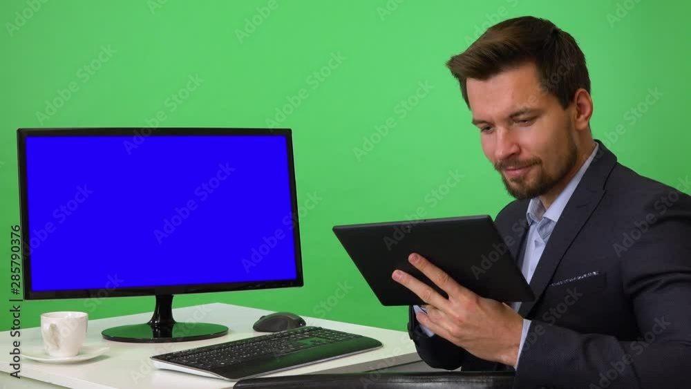 A young businessman sits in front of a computer with blue screen and works on a tablet, then smiles at the camera - green screen studio A young businessman sits in front of a computer with blue screen and works on a tablet, then smiles at the camera - green screen studio