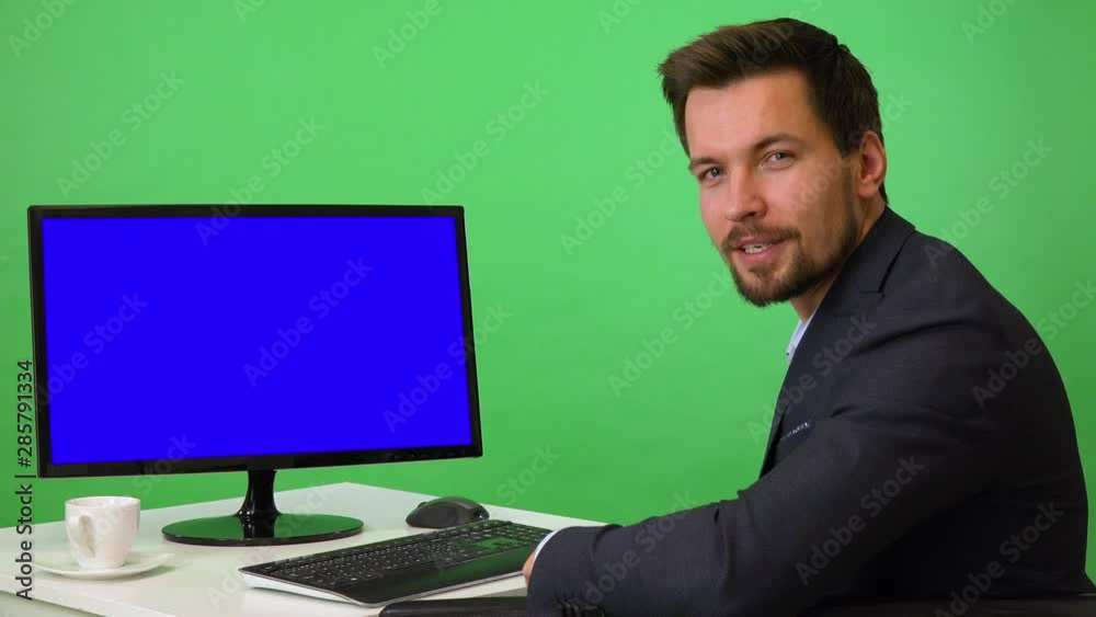 A young businessman sits in front of a computer with blue screen and talks to the camera with a smile - green screen studio A young businessman sits in front of a computer with blue screen and talks to the camera with a smile - green screen studio