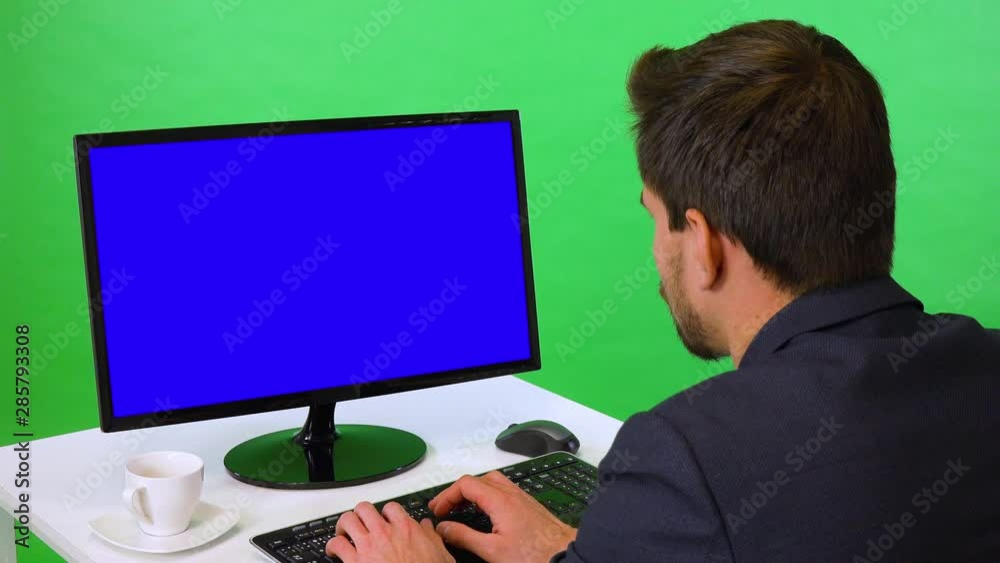 A young businessman sits at a desk and works on a desktop computer with blue screen - closeup - green screen studio - he types on keyboard A young businessman sits at a desk and works on a desktop computer with blue screen - closeup - green screen studio - he types on keyboard