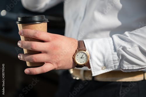 Young successful businessman in a white shirt drinking coffee standing near his luxary car. Driver takes a break to drink coffee.
