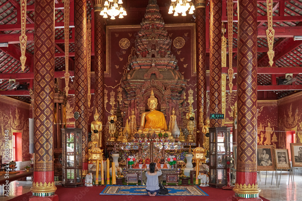 nside a Buddhist temple in Thailand, chaired by a large golden Buddha, in the middle there is a tourist on her knees who takes pictures