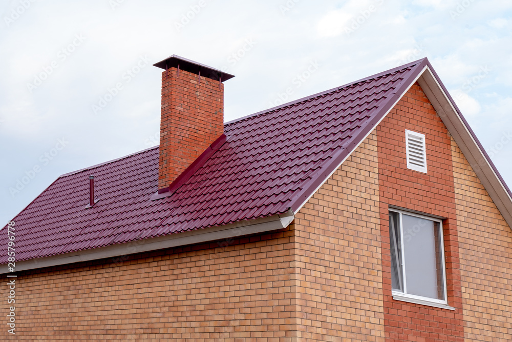 Part of a two-story private building of red brick against a clear sky. Fragment of a roof with a pipe and a window on the house. New affordable housing under the state program or mortgage lending.