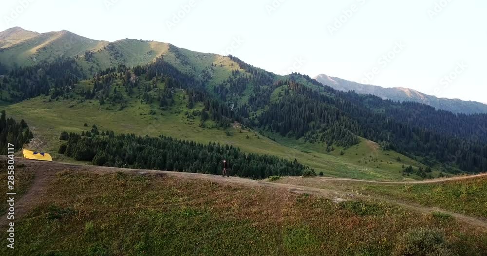 The guy enjoys the beauty of the surrounding nature. It offers views of green hills, tents, a guy on a hill, green coniferous trees and grass. Mountainous area. Tents are pitched on the hills. The guy enjoys the beauty of the surrounding nature. It offers views of green hills, tents, a guy on a hill, green coniferous trees and grass. Mountainous area. Tents are pitched on the hills.