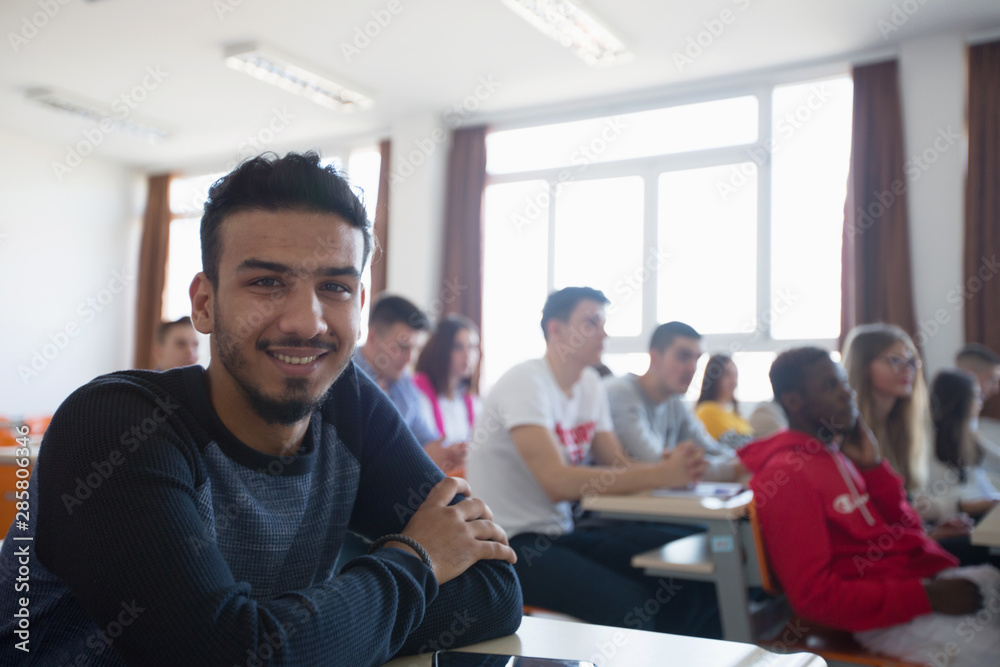 Smiling african american university male student in class sits at his desk turning to smile at the camera. University mixed race Students, african, american and european during the class. Smiling african american university male student in class sits at his desk turning to smile at the camera. University mixed race Students, african, american and european during the class.