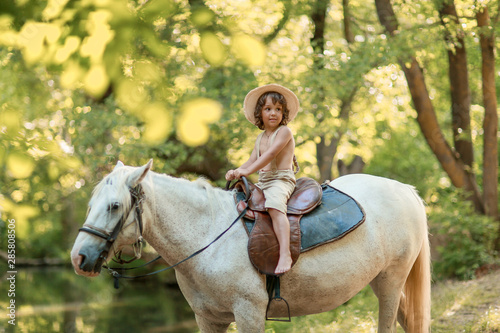 Little baby boy with curly hair dressed as hobbit playing with horse in summer forest