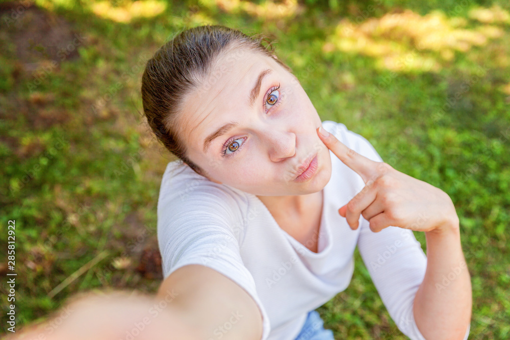 Young funny girl take selfie from hands with phone sitting on green grass park or garden background. Portrait of young attractive woman making selfie photo on smartphone in summer day Young funny girl take selfie from hands with phone sitting on green grass park or garden background. Portrait of young attractive woman making selfie photo on smartphone in summer day