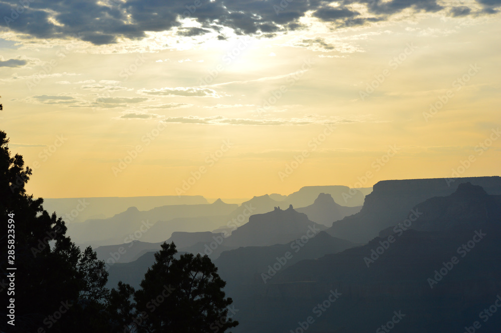 Landscape of the Grand Canyon ridges paled out as the sun peaks from behind a cloud, trees silhouettes in the foreground