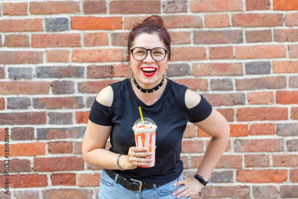 Fototapeta premium Cute cheerful young woman in stylish clothes is laughing and holding a milkshake in her hands against a brick wall. The concept of a summer holiday in the city. Copyspace.