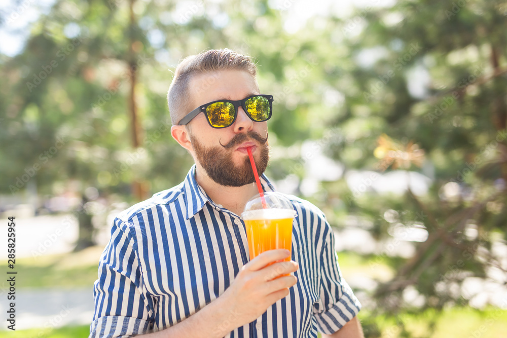 Fototapeta premium Portrait of a positive cheerful young man with a glass of juice with a straw while walking in the park on a warm sunny summer day. The concept of rest after study and work on weekends.