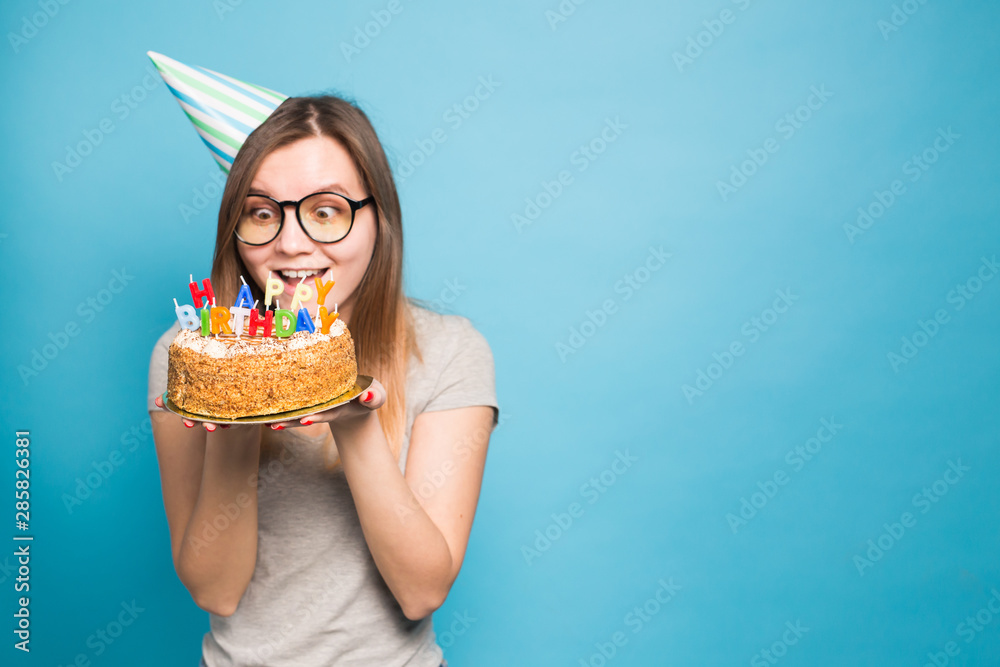 Crazy funny girl in a paper hat and glasses holding a big birthday cake on the blue background with copy space. Concept of prank and greetings.