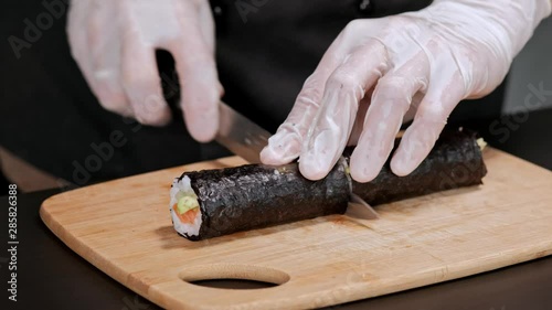 Young male sushi chef prepares Japanese sushi rolls of rice, salmon, avocado and nori. Restaurant kitchen, closeup hands cut a roll with a knife.