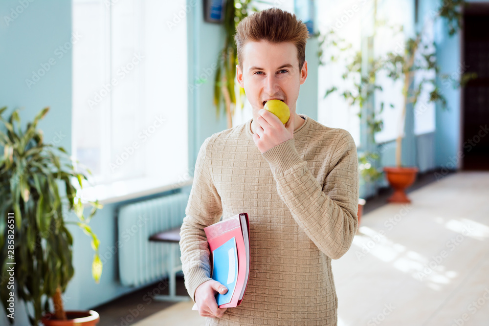 Happy male student eating an apple standing in university corridor while break between classes. Hardworking learner having healthy snack while studying Happy male student eating an apple standing in university corridor while break between classes. Hardworking learner having healthy snack while studying