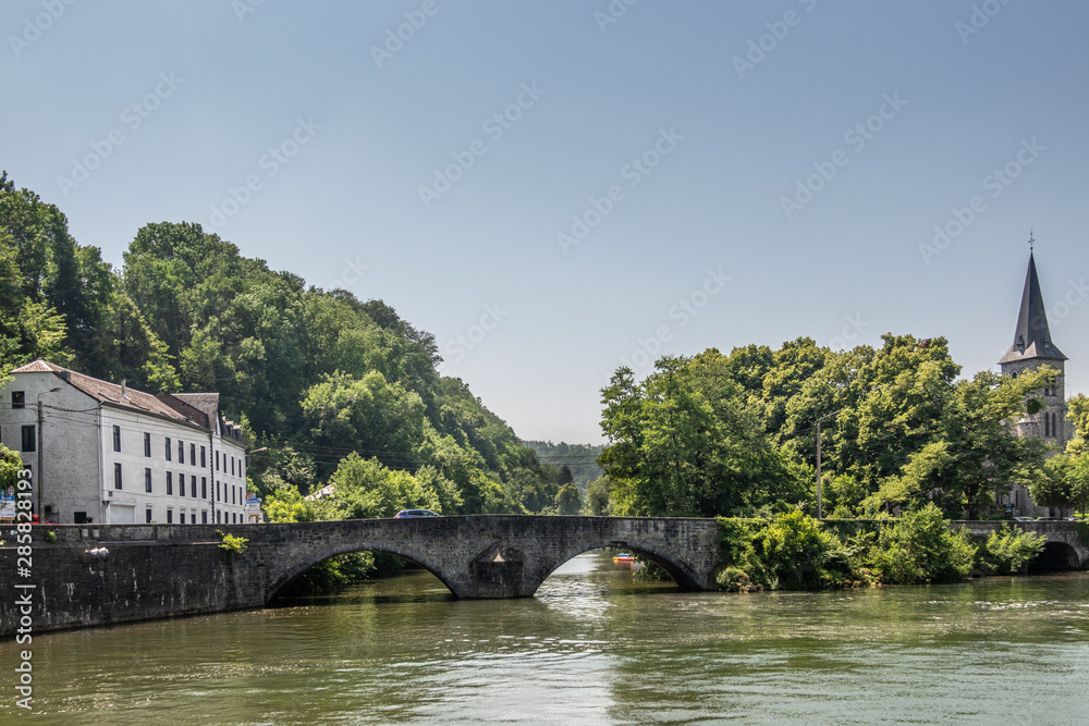 Fototapeta premium Dinant, Belgium - June 26, 2019: Old roman bridge over greenish Lesse River. Saint Anne Church tower captured in green foliage on the right. White building on the left under light blue sky.