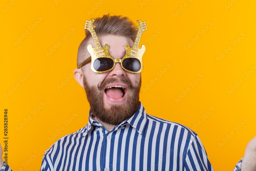 Positive young man with glasses in the form of guitars rejoices against a yellow background. The concept of celebration and parties.
