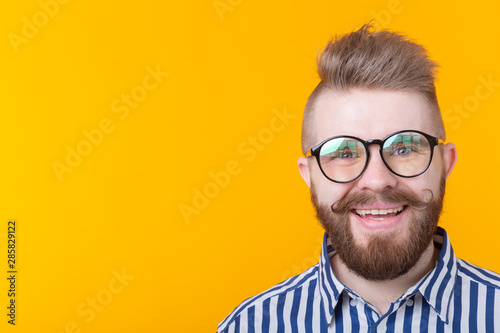 Young positive trendy man hipster with a mustache beard and fetish necklace in shirt posing on a yellow background with copy space. Concept of rock and subculture.