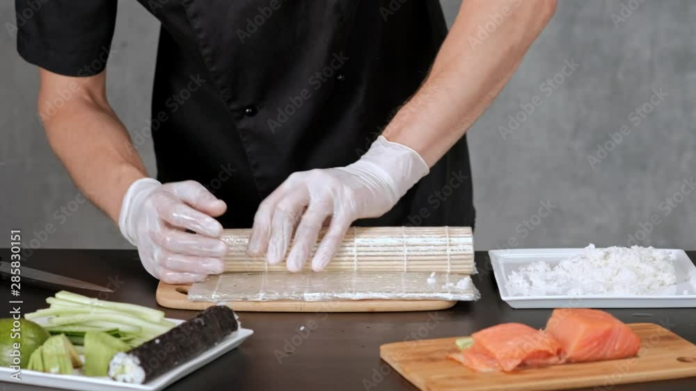 Young male sushi chef prepares Japanese sushi rolls of rice, salmon, avocado and nori. Restaurant kitchen, hands closeup, rolls on mat. Young male sushi chef prepares Japanese sushi rolls of rice, salmon, avocado and nori. Restaurant kitchen, hands closeup, rolls on mat.