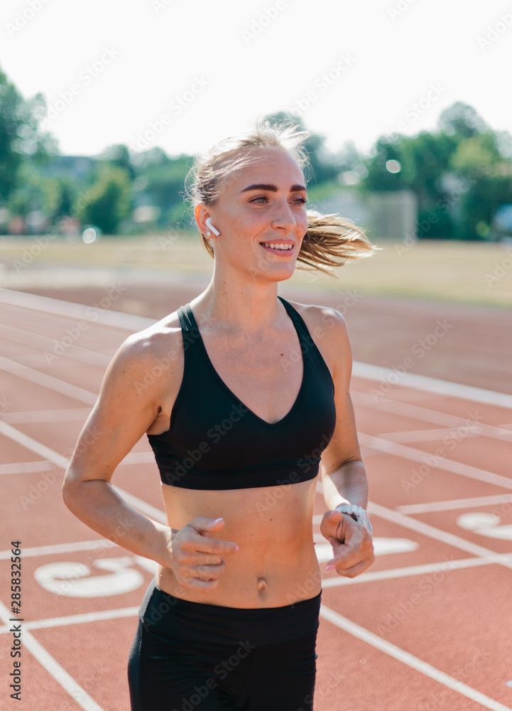 Young cheerful woman runner in sportswear running on stadium track with red coating outdoors Young cheerful woman runner in sportswear running on stadium track with red coating outdoors