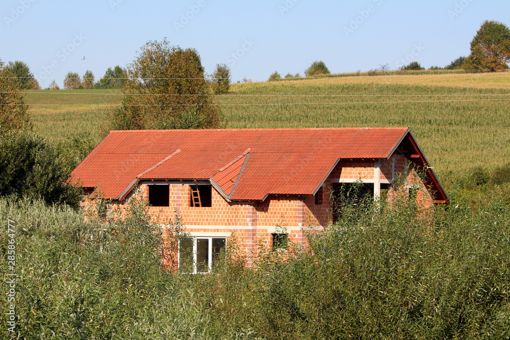 Large new unfinished red brick family house still under construction with new roof tiles and without windows completely surrounded with tall grass and forest vegetation on warm sunny summer day