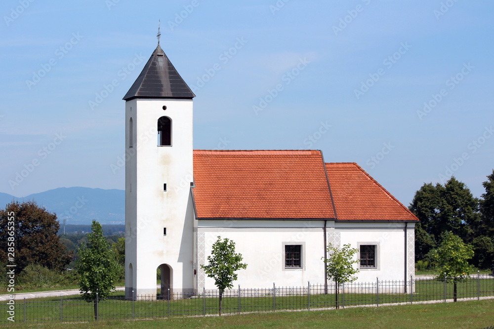 Local country church with white facade and new roof tiles surrounded with metal fence and freshly cut grass on clear blue sky background