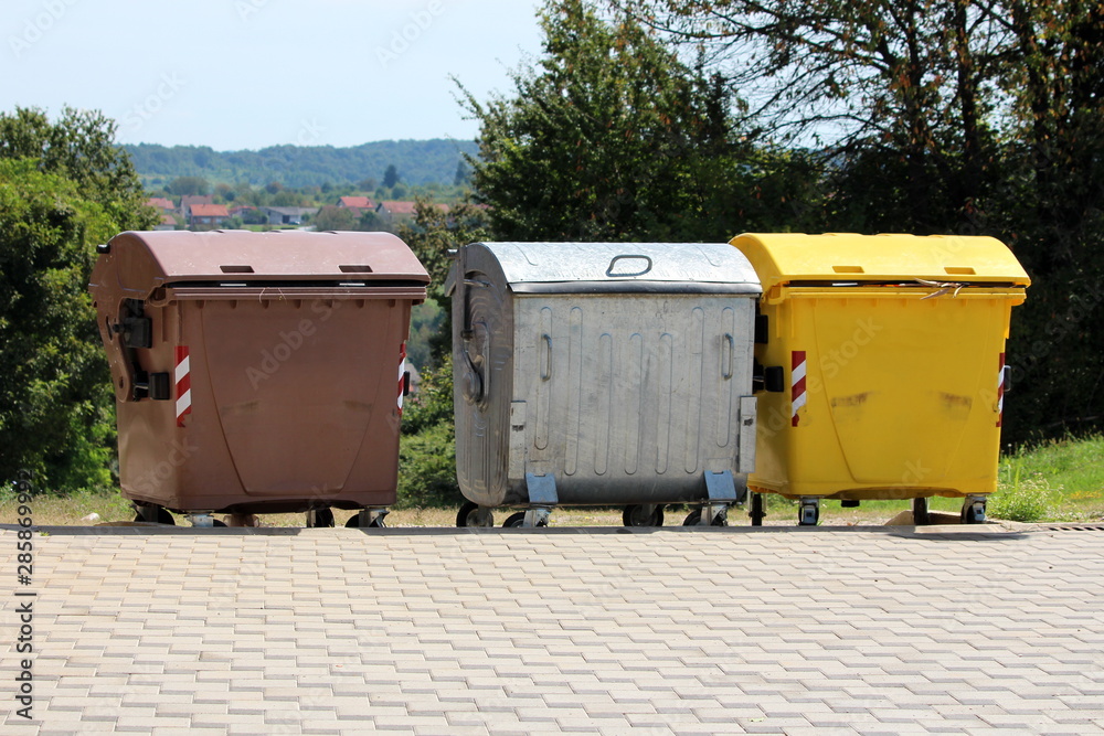 Three various large metal and plastic trash containers for ecologically sorting garbage for recycling surrounded with stone tiles and trees on warm sunny summer day Three various large metal and plastic trash containers for ecologically sorting garbage for recycling surrounded with stone tiles and trees on warm sunny summer day