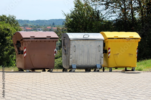 Three various large metal and plastic trash containers for ecologically sorting garbage for recycling surrounded with stone tiles and trees on warm sunny summer day